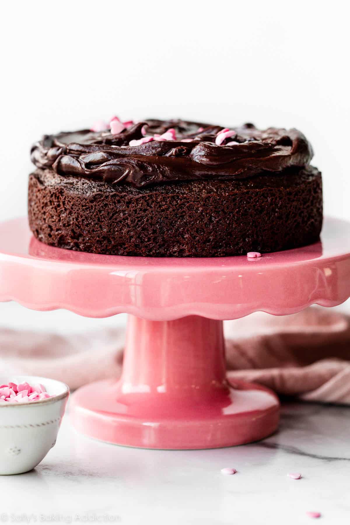 6 inch chocolate cake with chocolate ganache on top sitting on a pink cake stand.