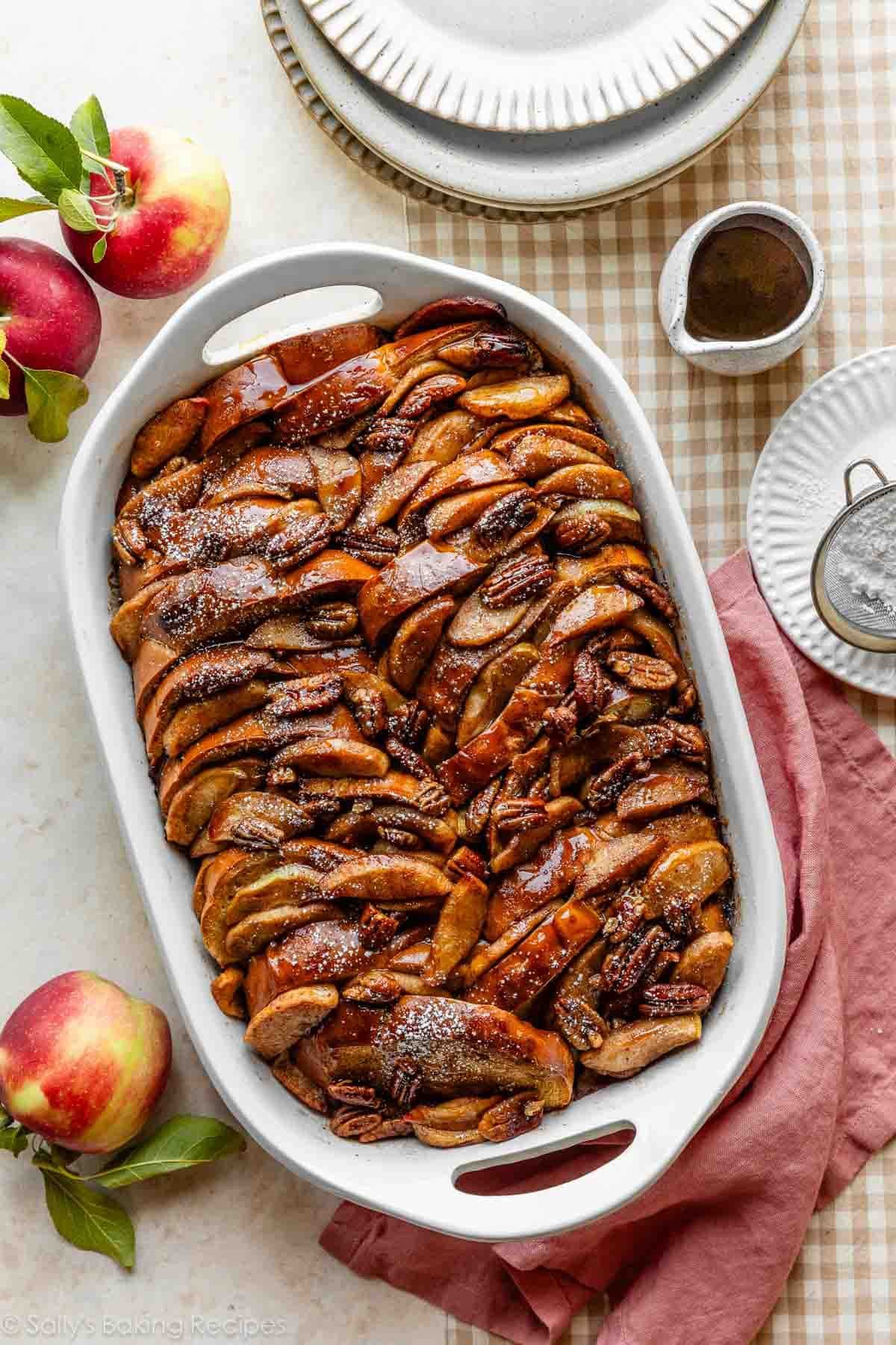 baked apple cider french toast casserole with pecans and maple syrup in white casserole dish with fresh apples.