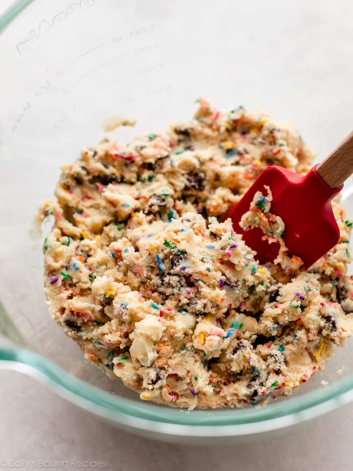 sprinkle chocolate chip cookie dough in glass bowl with red silicone spatula.