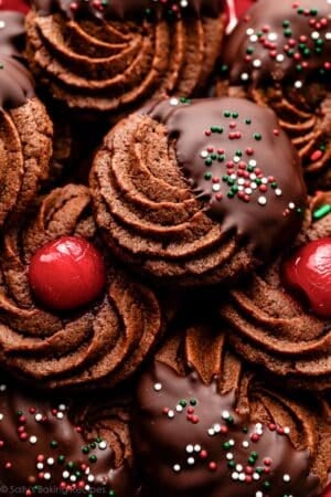 plate of chocolate butter cookies dipped in chocolate with sprinkles and maraschino cherries.