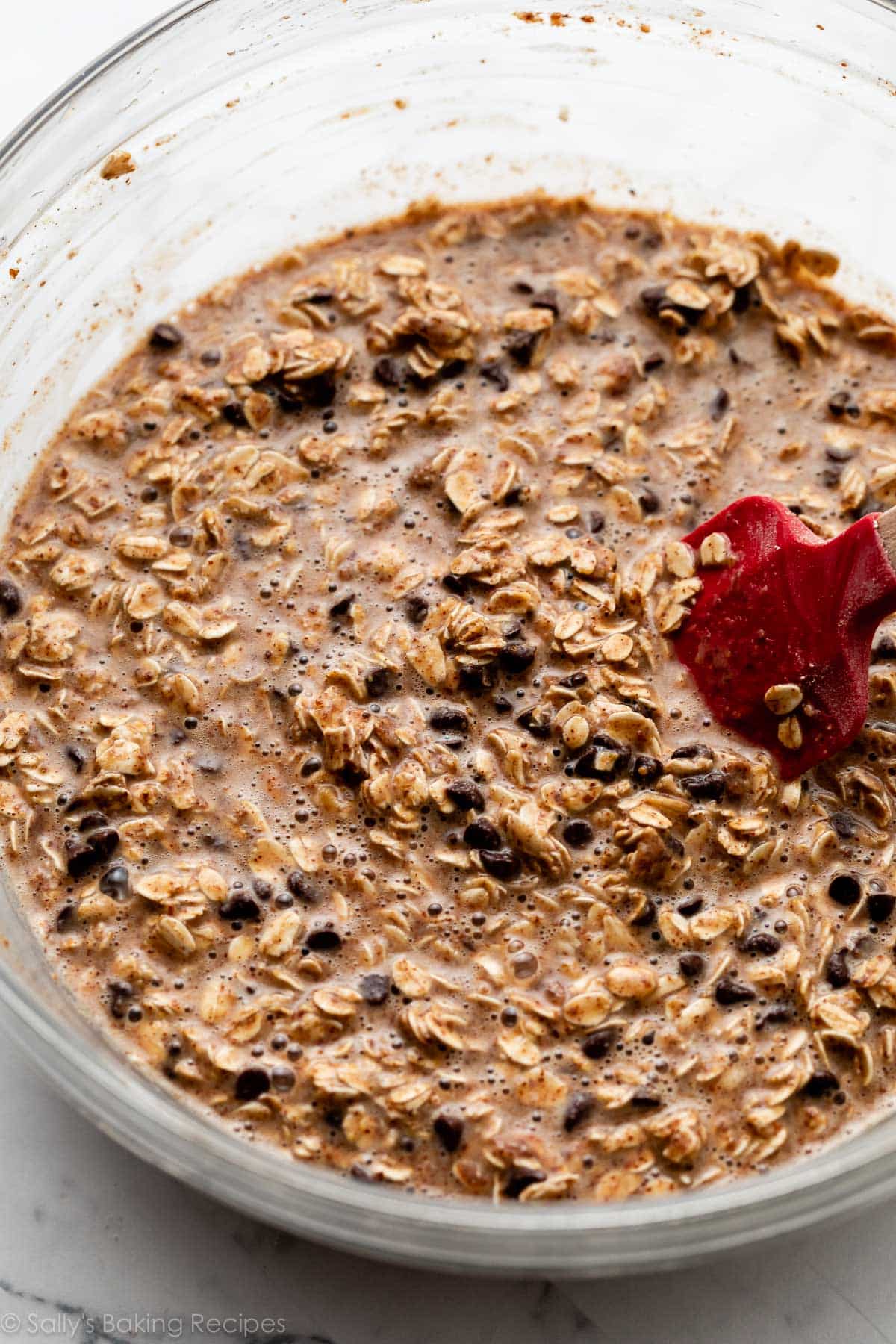 oats in liquid batter with mini chocolate chips in a glass bowl with red silicone spatula.