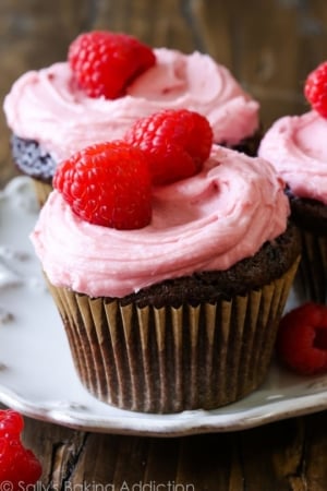 chocolate cupcakes topped with raspberry frosting, sprinkles, and raspberries on a white plate