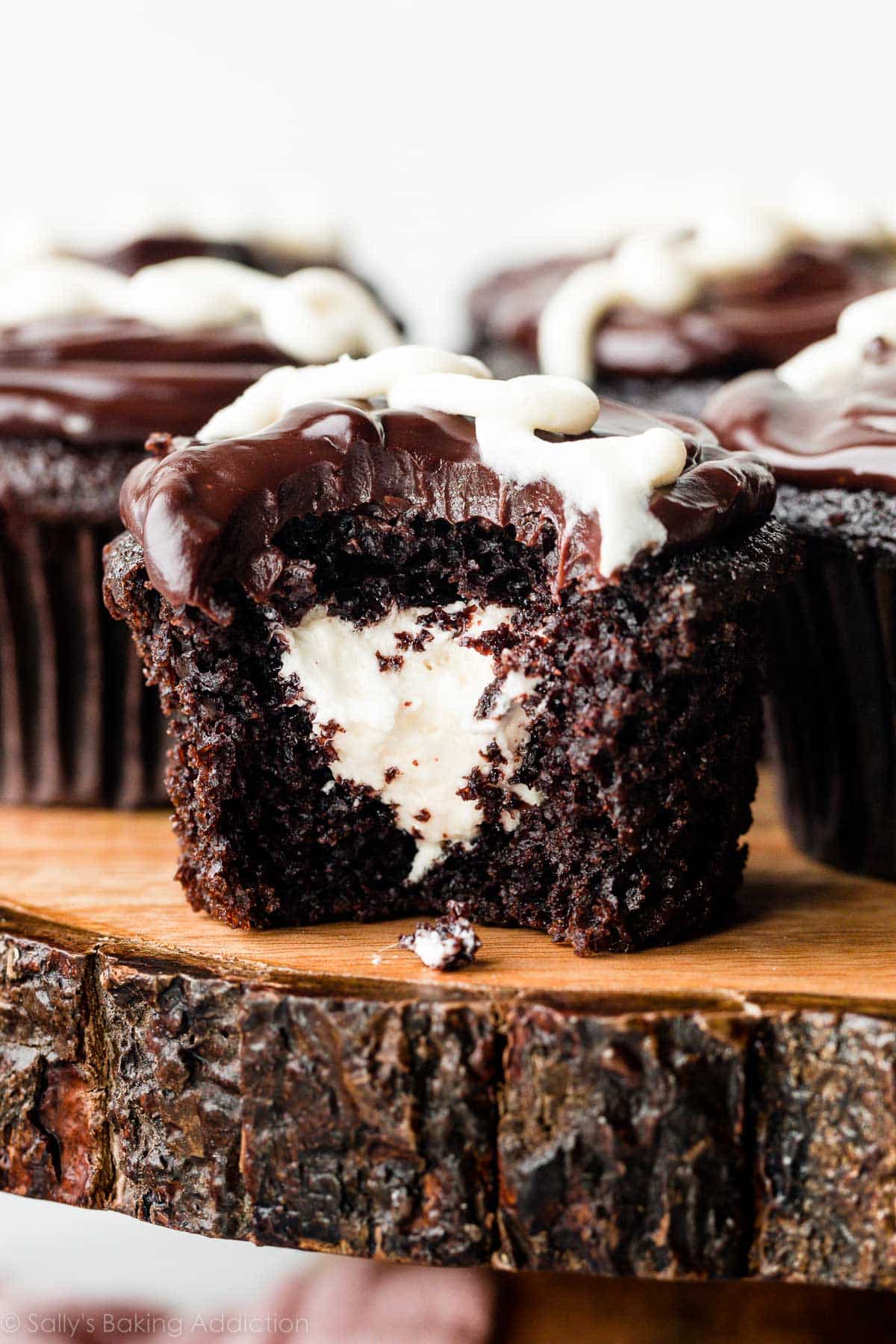 cream-filled chocolate cupcakes on wooden cake stand and 1 cupcake has bite taken out to reveal cream center.