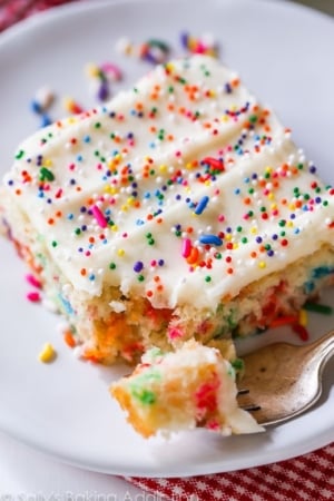 slice of funfetti sheet cake topped with vanilla frosting and sprinkles on a white plate with a fork