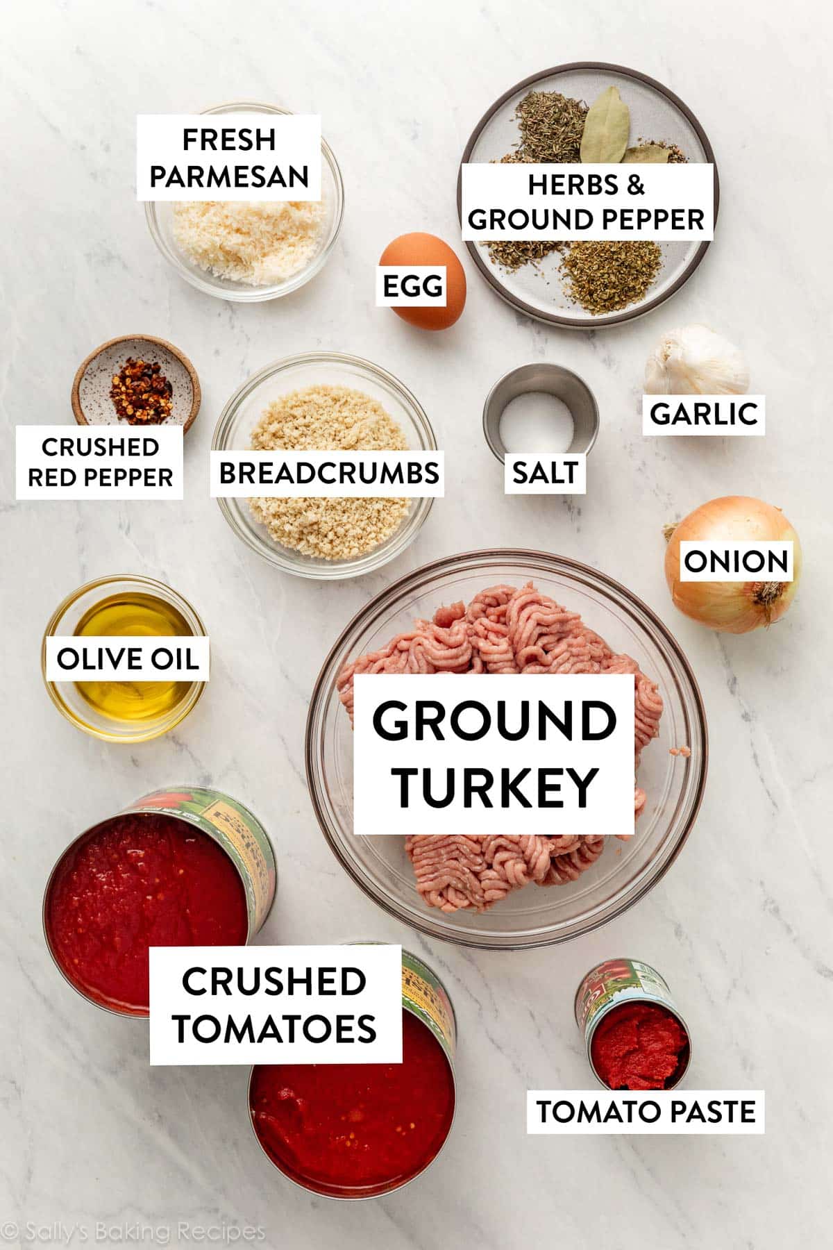 ingredients measured out on marble surface including herbs, garlic, onion, breadcrumbs, cans of crushed tomatoes and tomato paste, and olive oil.