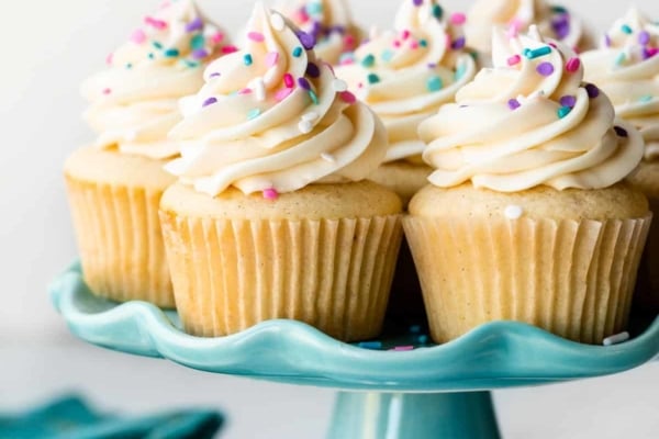 vanilla cupcakes on blue cake stand with pink, purple, and blue sprinkles on top.