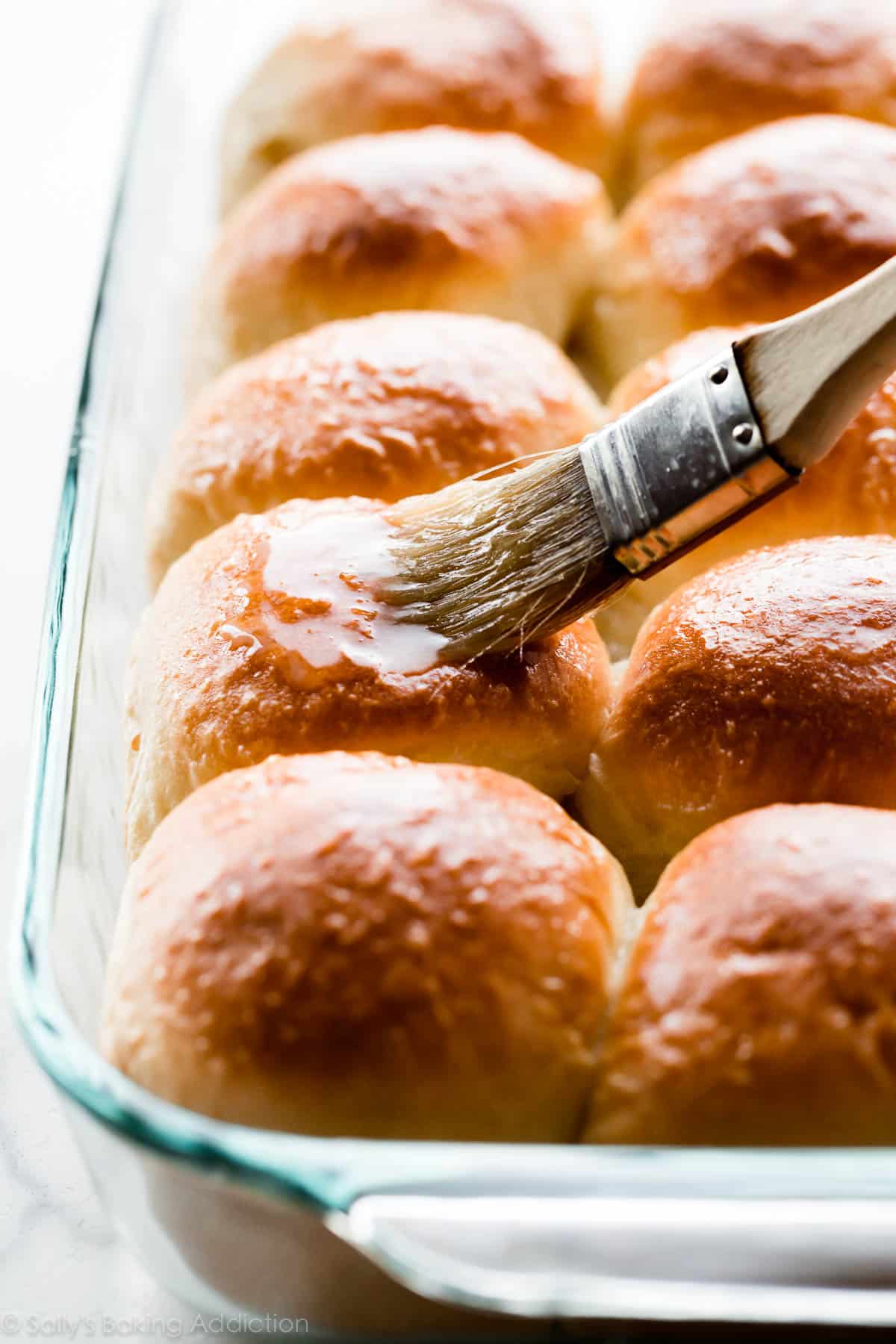 brushing honey butter topping onto dinner rolls in a glass baking pan