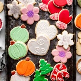 decorated sugar cookies on baking sheet including hearts, pumpkins, trees, mittens, and flowers.