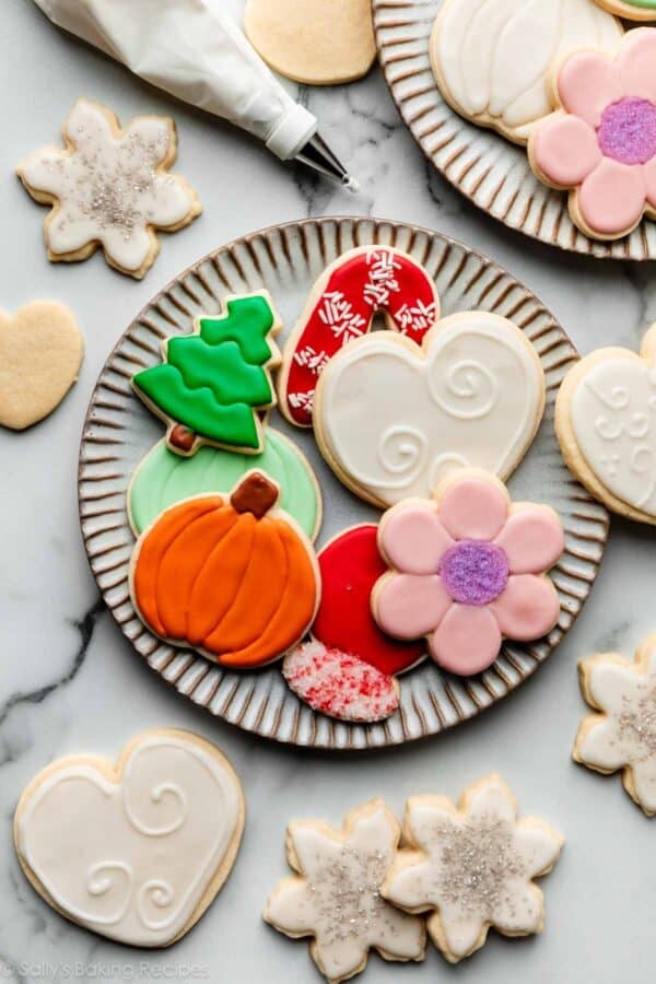 various shaped decorated sugar cookies on plate including a pink iced flower, orange pumpkin, and a green tree cookie.