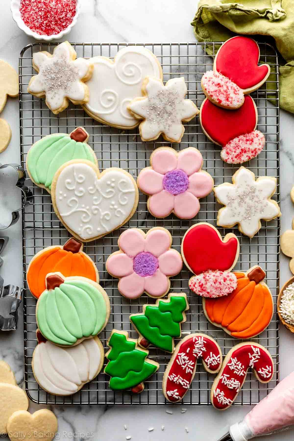 Decorated sugar cookies including pink flowers, white hearts, red mittens, orange pumpkins, and green trees on wire cooling rack.