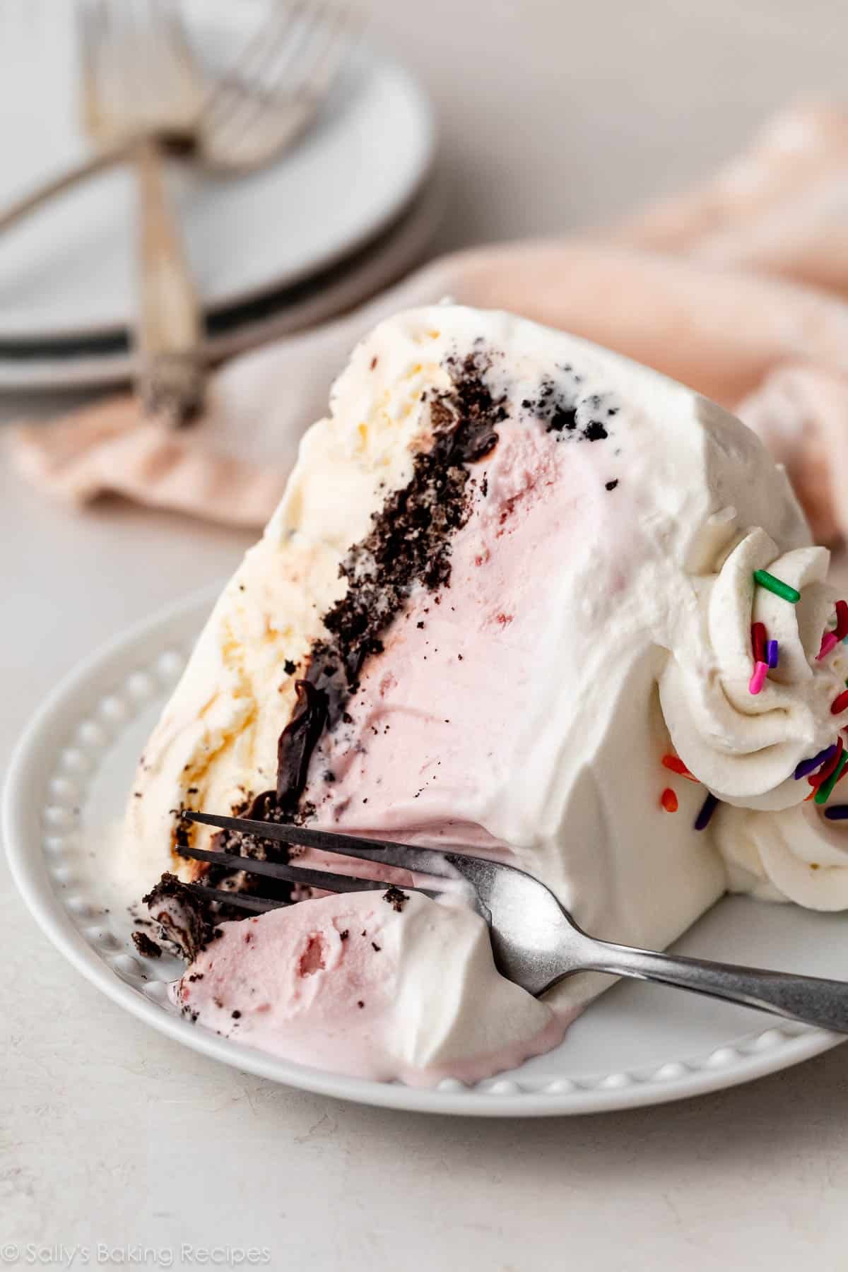 vanilla, Oreo, and strawberry ice cream cake with whipped cream on white plate with fork.