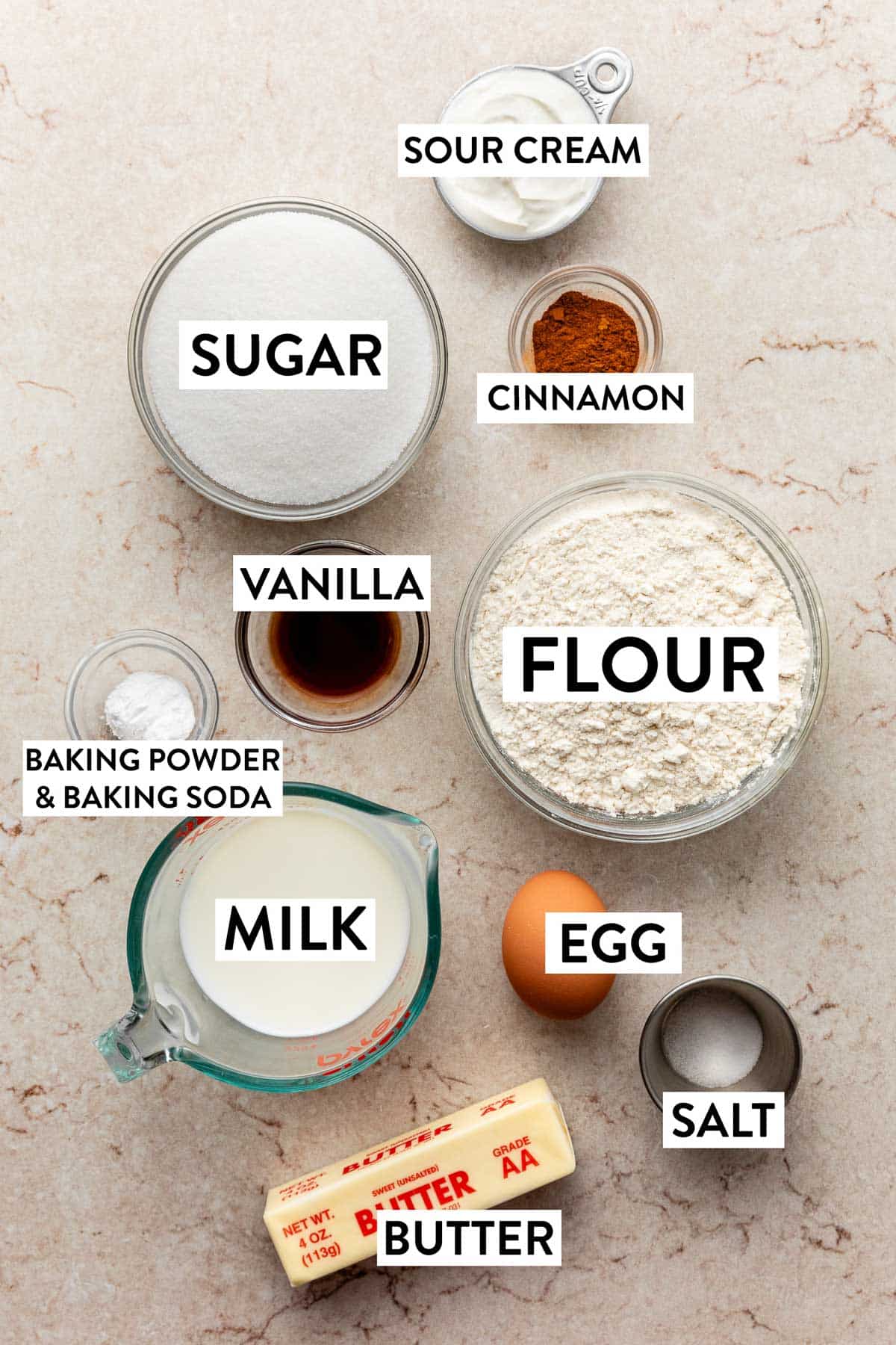 ingredients on marbled surface including flour, egg, milk, butter, salt, vanilla, and sugar.