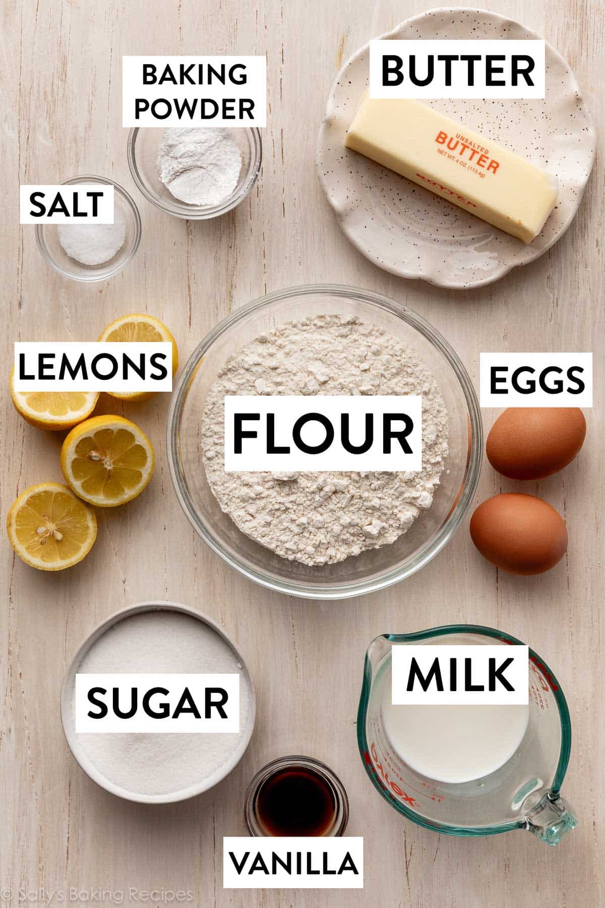 ingredients on wooden backdrop including flour, butter, eggs, milk, sugar, and vanilla.