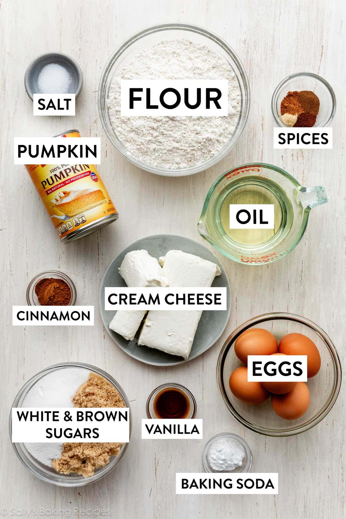 ingredients on white wooden surface including oil, pumpkin, flour, eggs, white sugar, brown sugar, and cinnamon.