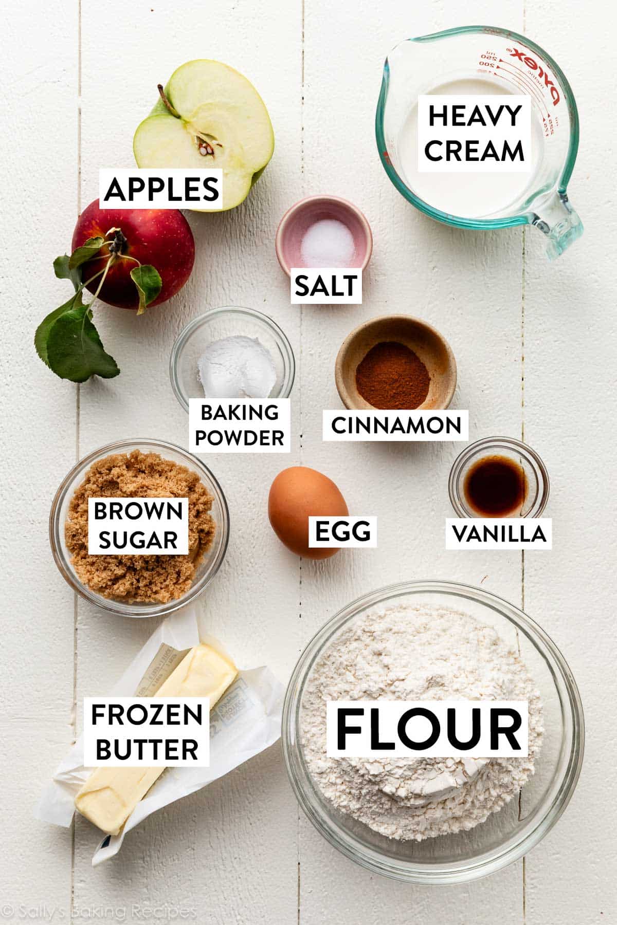 ingredients on white wooden backdrop including flour, butter, heavy cream, apple, brown sugar, and egg.