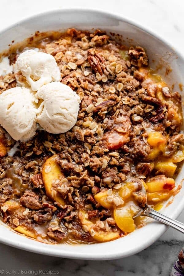 peach pecan crisp in white round baking dish with 3 vanilla ice cream scoops on top.