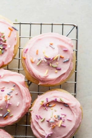 sugar cookies topped with pink buttercream and sprinkles on a cooling rack