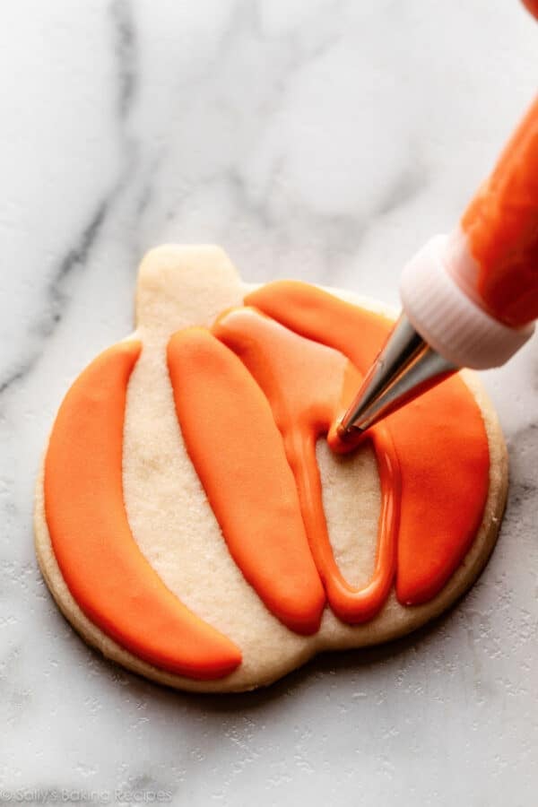 piping orange icing decoration on pumpkin-shaped cookie.