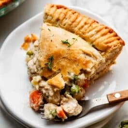 slice of double crust chicken pot pie with golden flaky pie crust on white plate with fork that has a wooden handle.