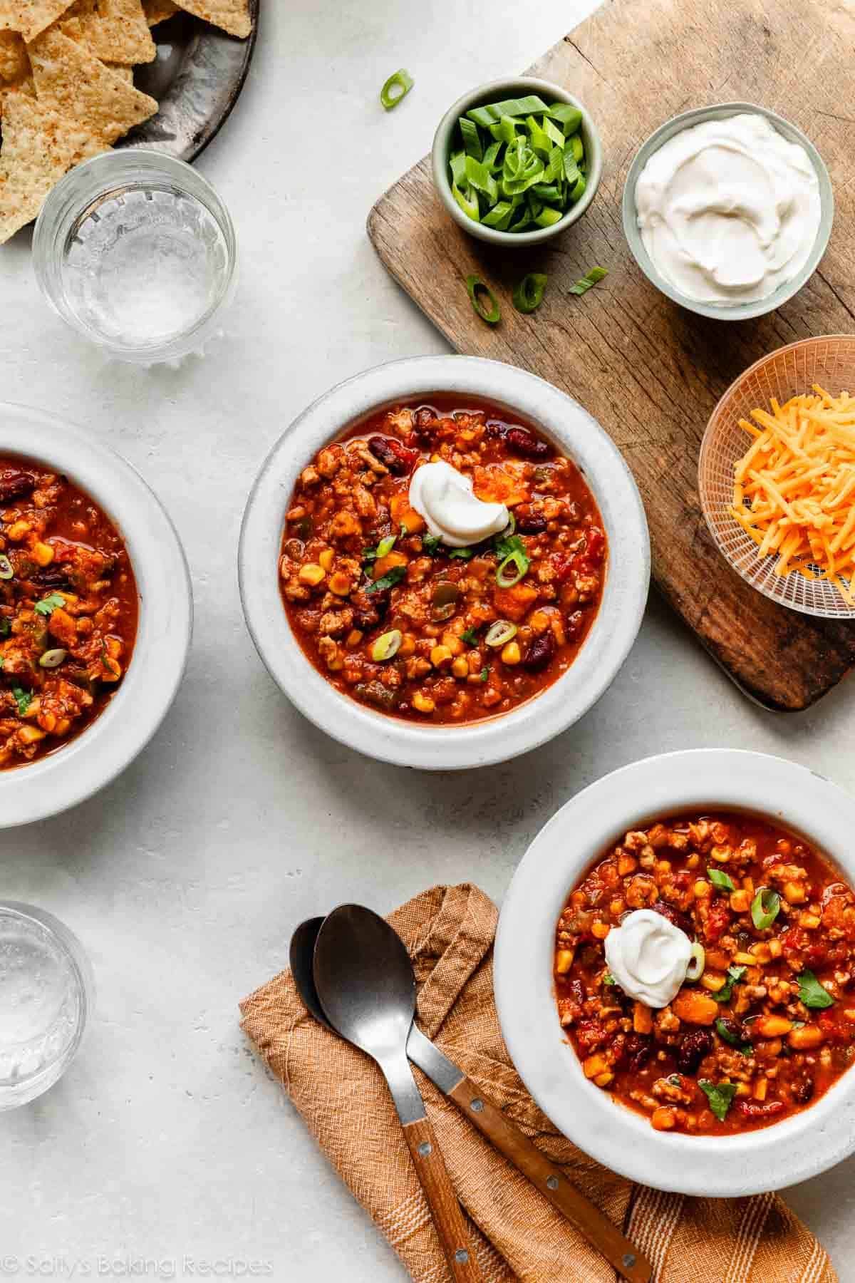 turkey chili in white bowls with smaller bowls of green onions, sour cream, and cheddar cheese near it.