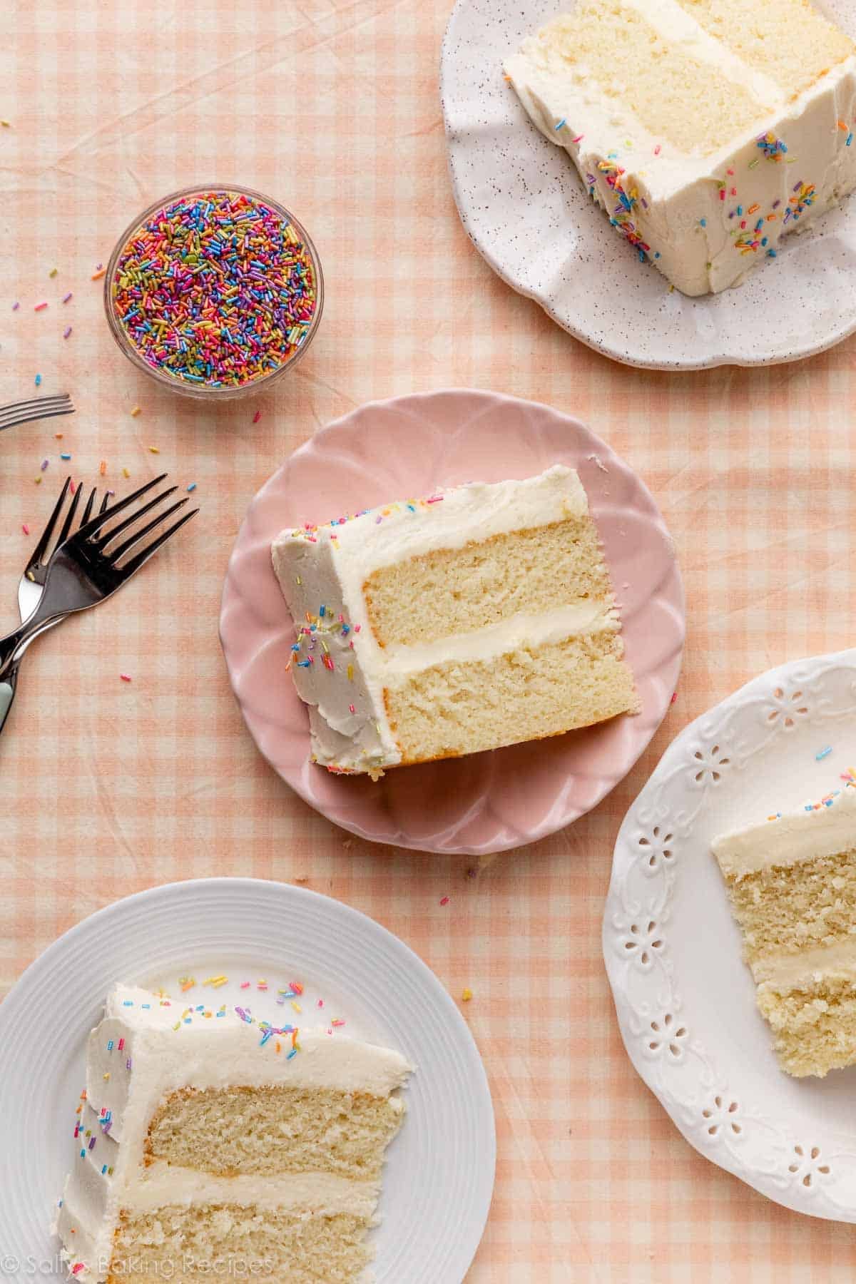 slices of white cake with vanilla frosting on assorted plates on light pink gingham linen.