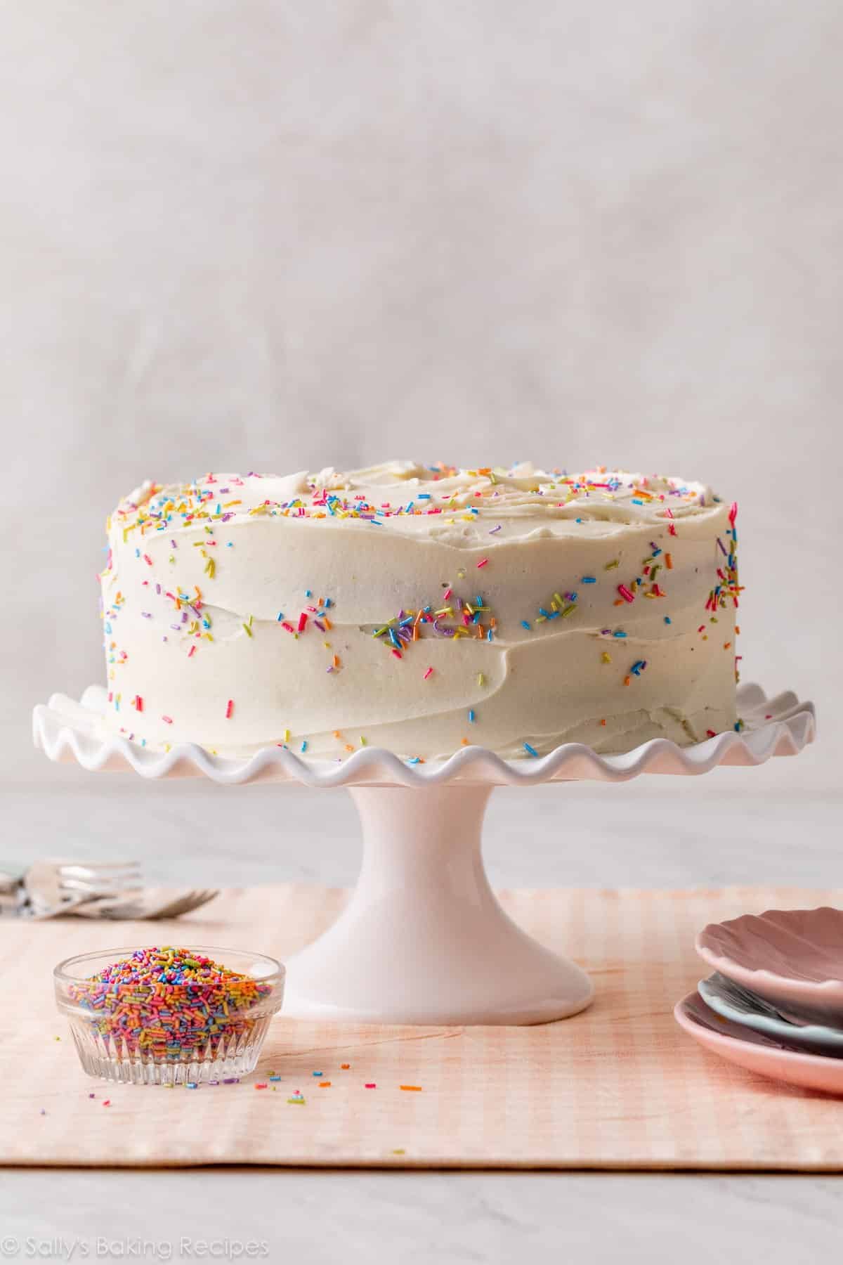 vanilla frosted cake with rainbow sprinkles on white ruffled cake stand.