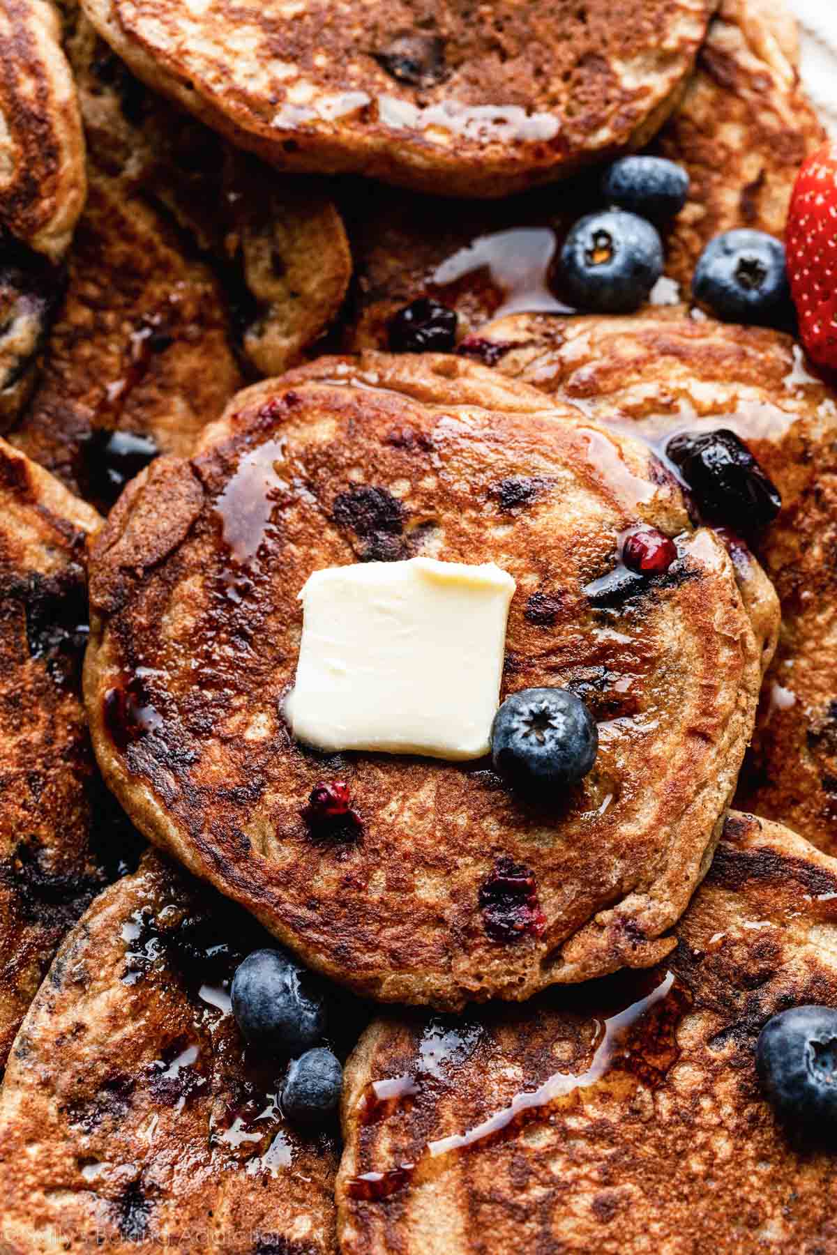 close-up of whole wheat blueberry pancakes with maple syrup, butter, and fresh blueberries on top.